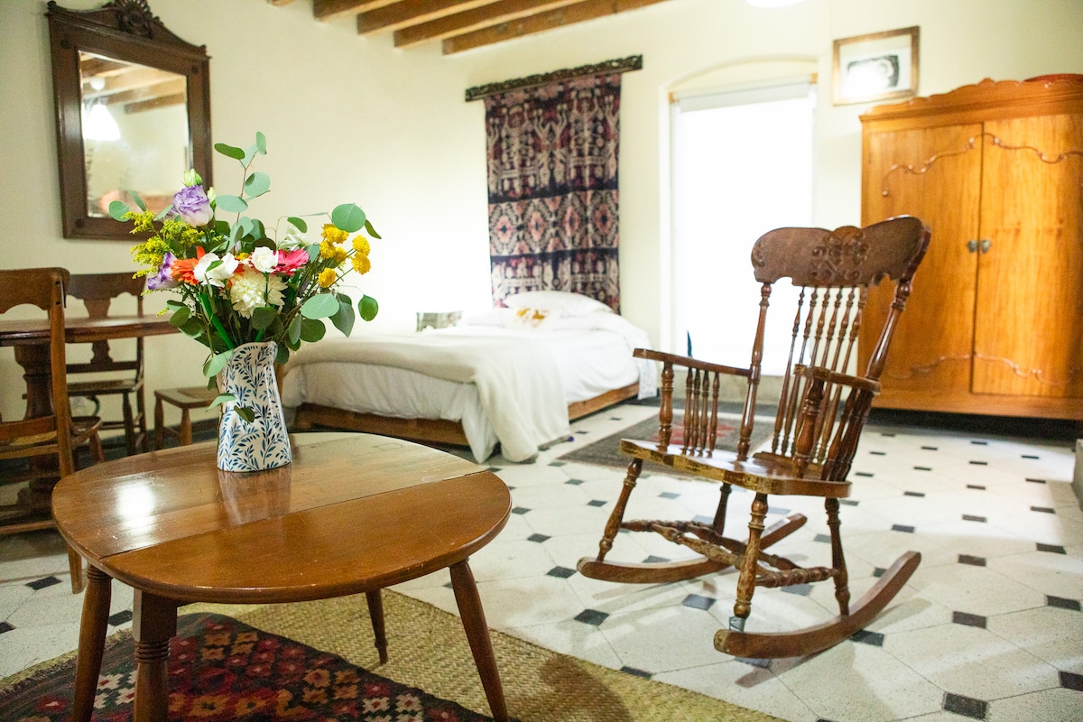 A charming room features a cozy rocking chair and a wooden table with a vase of fresh flowers. Soft light fills the space, highlighting the spacious bed and an intricately designed armoire. The floor is adorned with patterned tiles, adding character to the setting.
