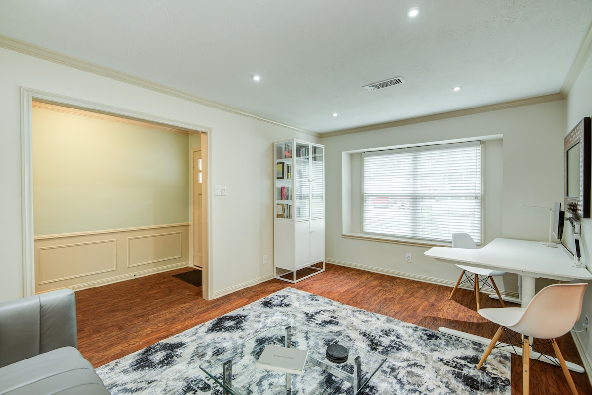 A well-lit living area features laminate wood flooring and large windows allowing natural light to fill the space. A plush area rug centers the room, while a small desk with a chair is positioned next to a storage unit, offering a functional workspace.