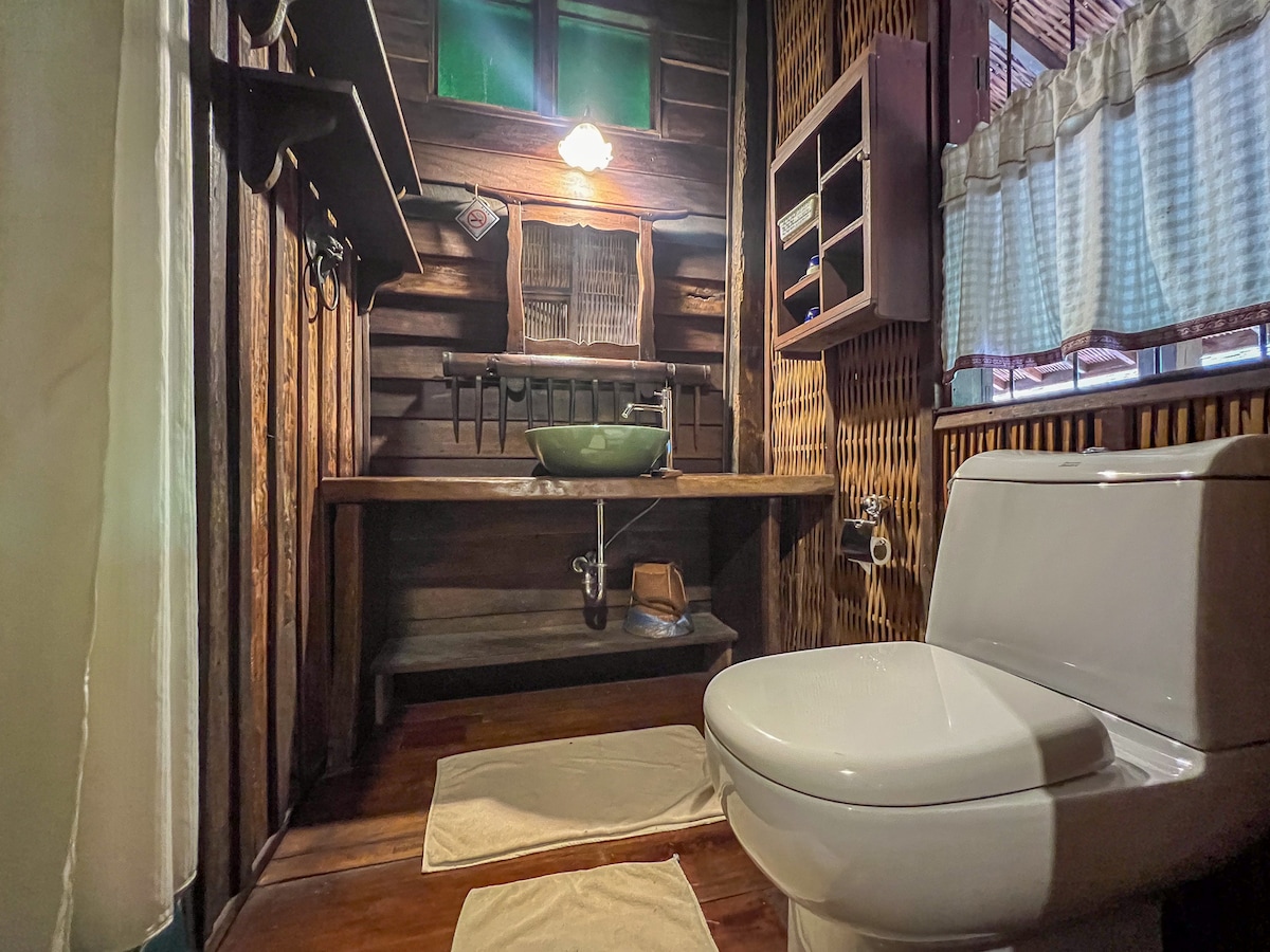 A rustic bathroom features wooden walls and a sink made from natural materials. A modern toilet is positioned beside the elegantly crafted storage shelves. Soft towels are placed on the floor, and gentle light filters in through a window covered by sheer curtains.