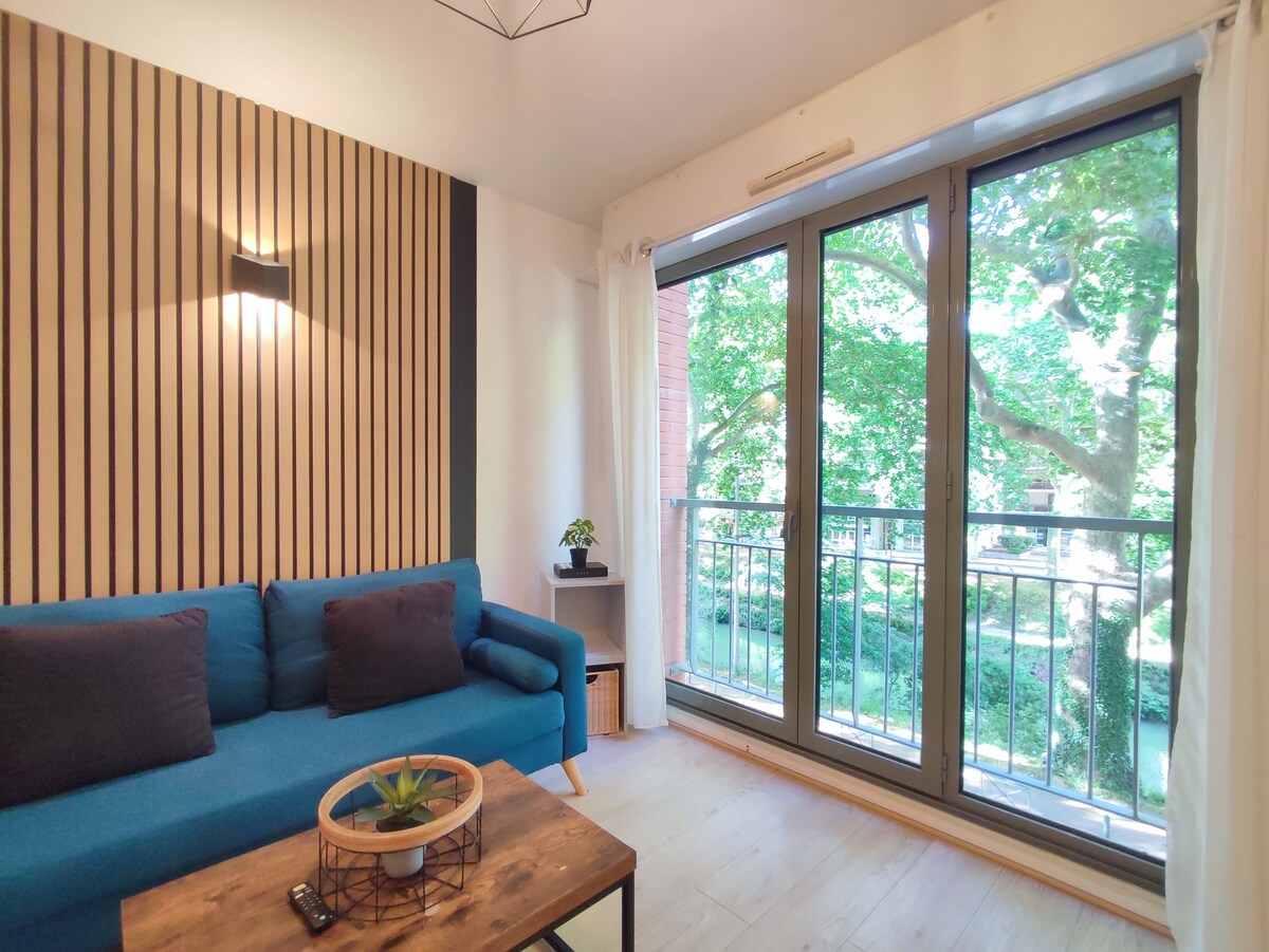 A modern seating area features a blue sofa with two dark cushions, positioned alongside a wooden coffee table. Large glass doors open to a balcony overlooking greenery, while natural light fills the space through sheer white curtains.