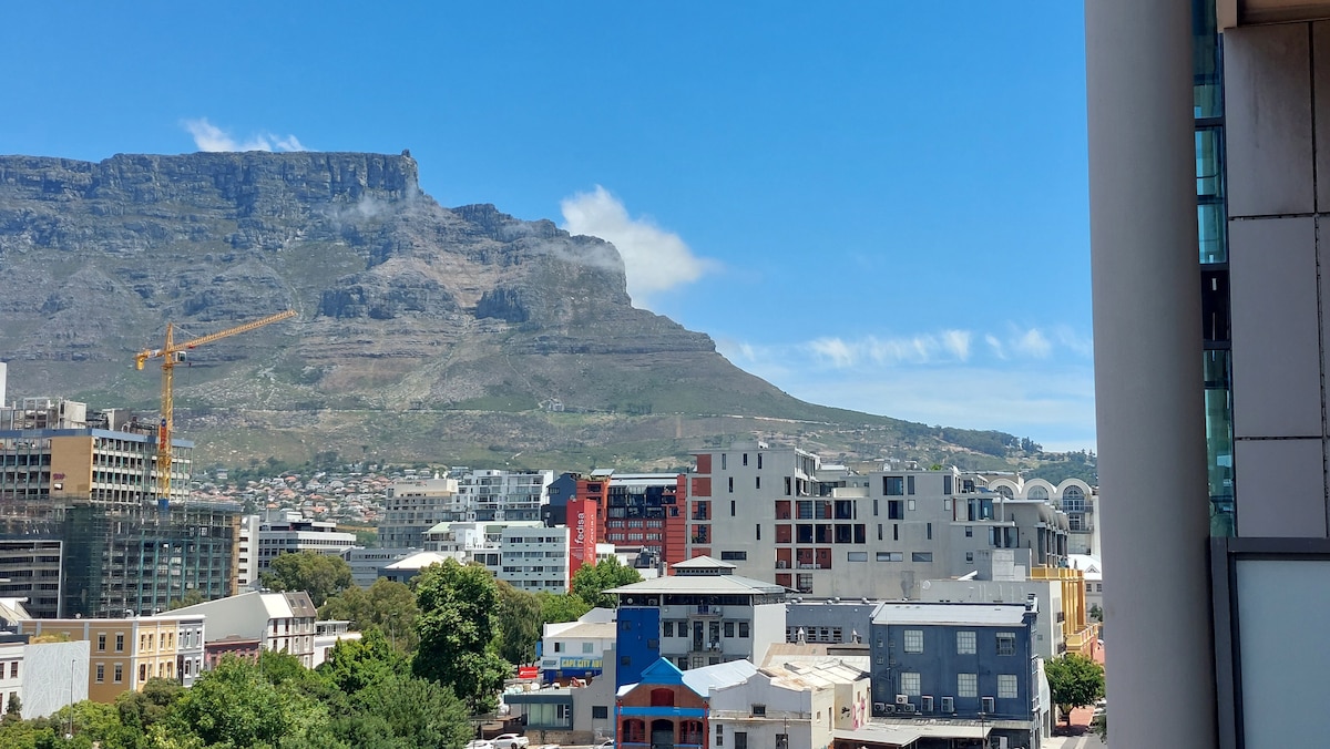A panoramic view showcases Table Mountain rising in the distance, framed by a blue sky. Various modern buildings and a construction crane are visible in the foreground, highlighting the vibrant urban environment of the area.
