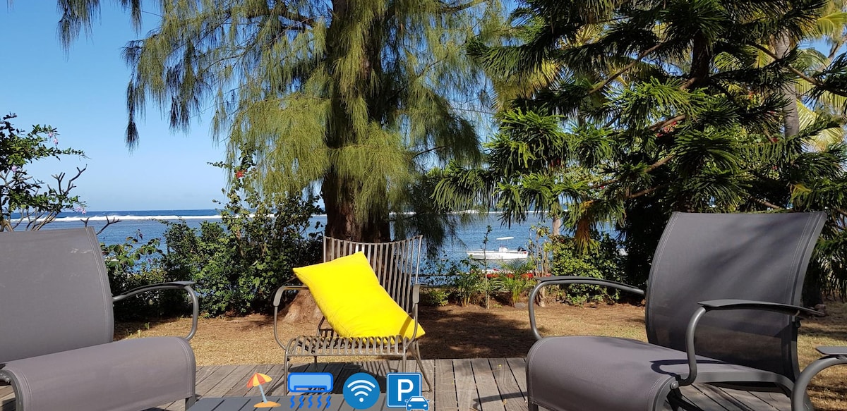 Two chairs and a stylish lounge chair are positioned on a wooden deck, offering a view of the lagoon in the background. Tropical plants surround the area, creating a serene outdoor space bathed in sunlight.