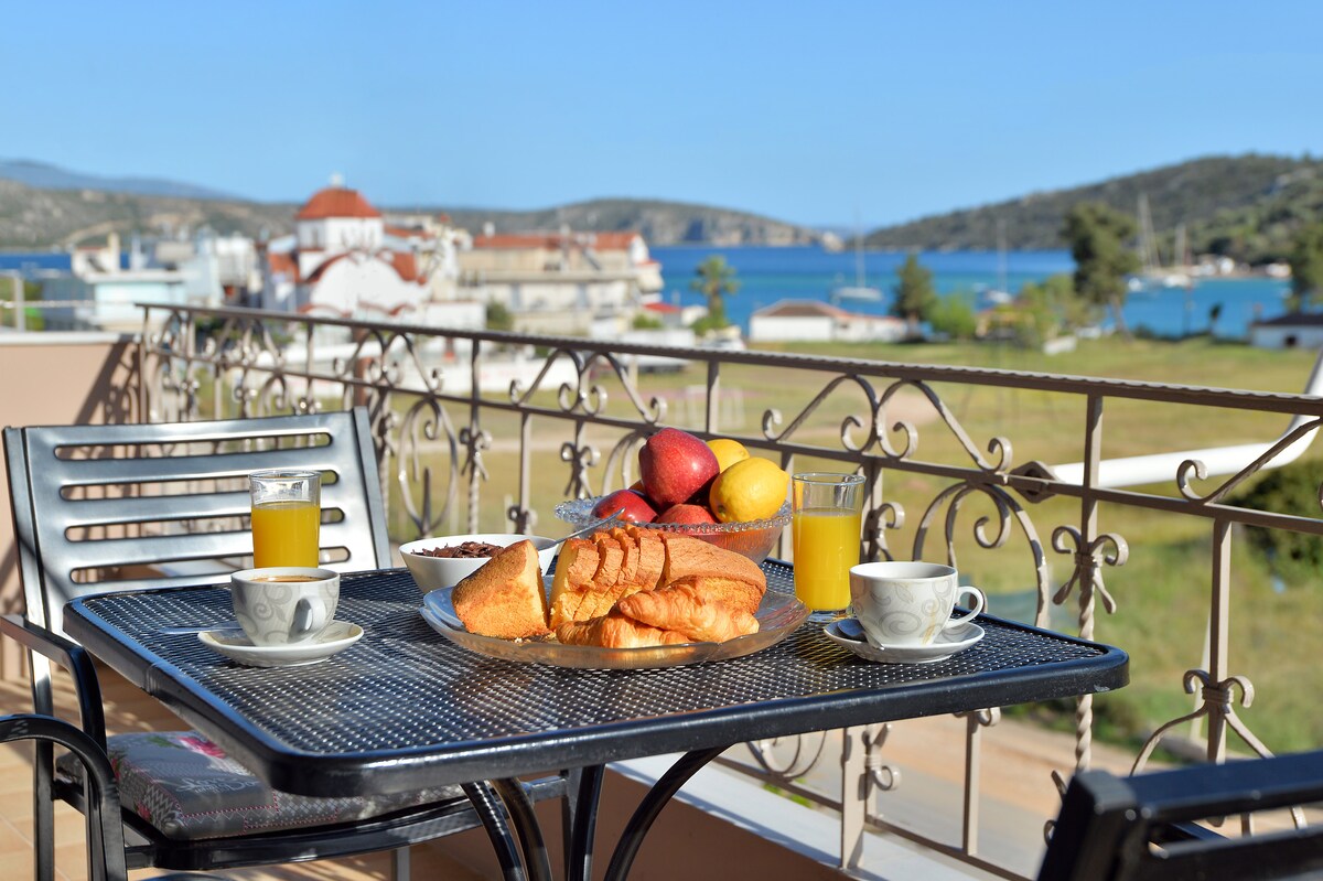 A private balcony is presented with a black metal table and four chairs. Breakfast items, including pastries and fruit, are arranged on a plate, accompanied by two cups and drinks. A serene sea view and coastal landscape complete the inviting outdoor setting.