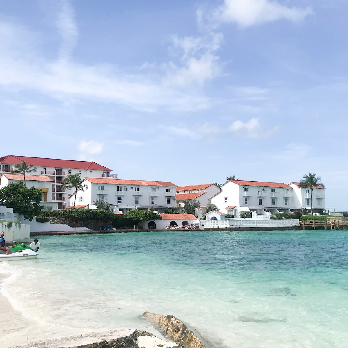 A white-walled resort complex is visible along the shoreline, featuring multiple buildings with red-tiled roofs. The clear turquoise water laps gently at the sandy beach, while palm trees add greenery to the landscape, enhancing the inviting coastal setting.