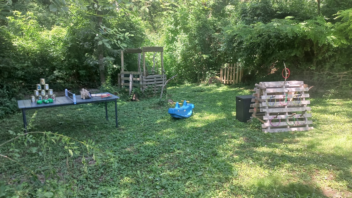 A green outdoor area features a grassy space surrounded by dense foliage. On the left, a table with various items is set up alongside a wooden play structure. To the right, a blue children's toy and a structure made of wooden pallets are visible, creating a playful atmosphere.