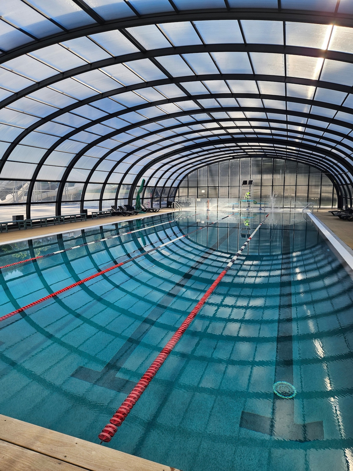 An indoor swimming pool is enclosed by a curved, transparent structure, allowing natural light to illuminate the water surface. Red lane markers are visible in the inviting blue water, with swimming lanes clearly defined. A view of distant poolside features can be seen through the arched ceiling.