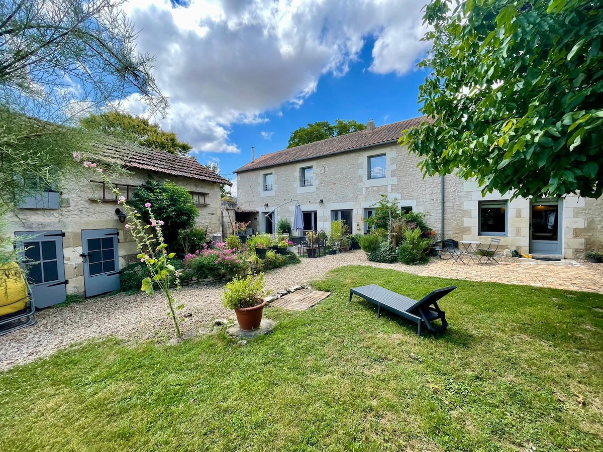 The shared garden area features well-maintained grass and a variety of plants, with lounging furniture placed among flowering shrubbery. Two sections of the longère are visible, showcasing their stone facades under a partly cloudy sky. Pathways connect the living areas to the garden.