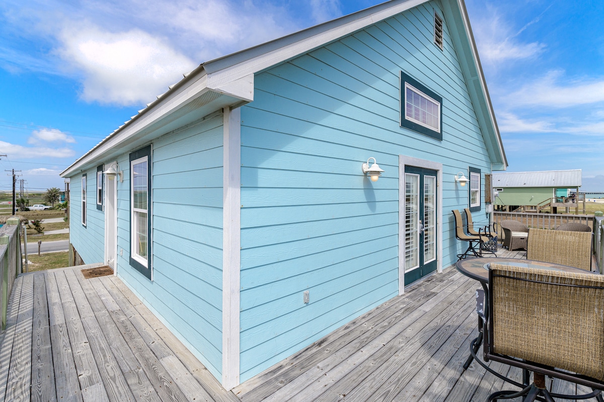 A light blue exterior of the beach house is shown, featuring a wide deck with seating. The facade includes large windows and double doors, allowing natural light to illuminate the interior. The surrounding area is a blend of grass and pavement.