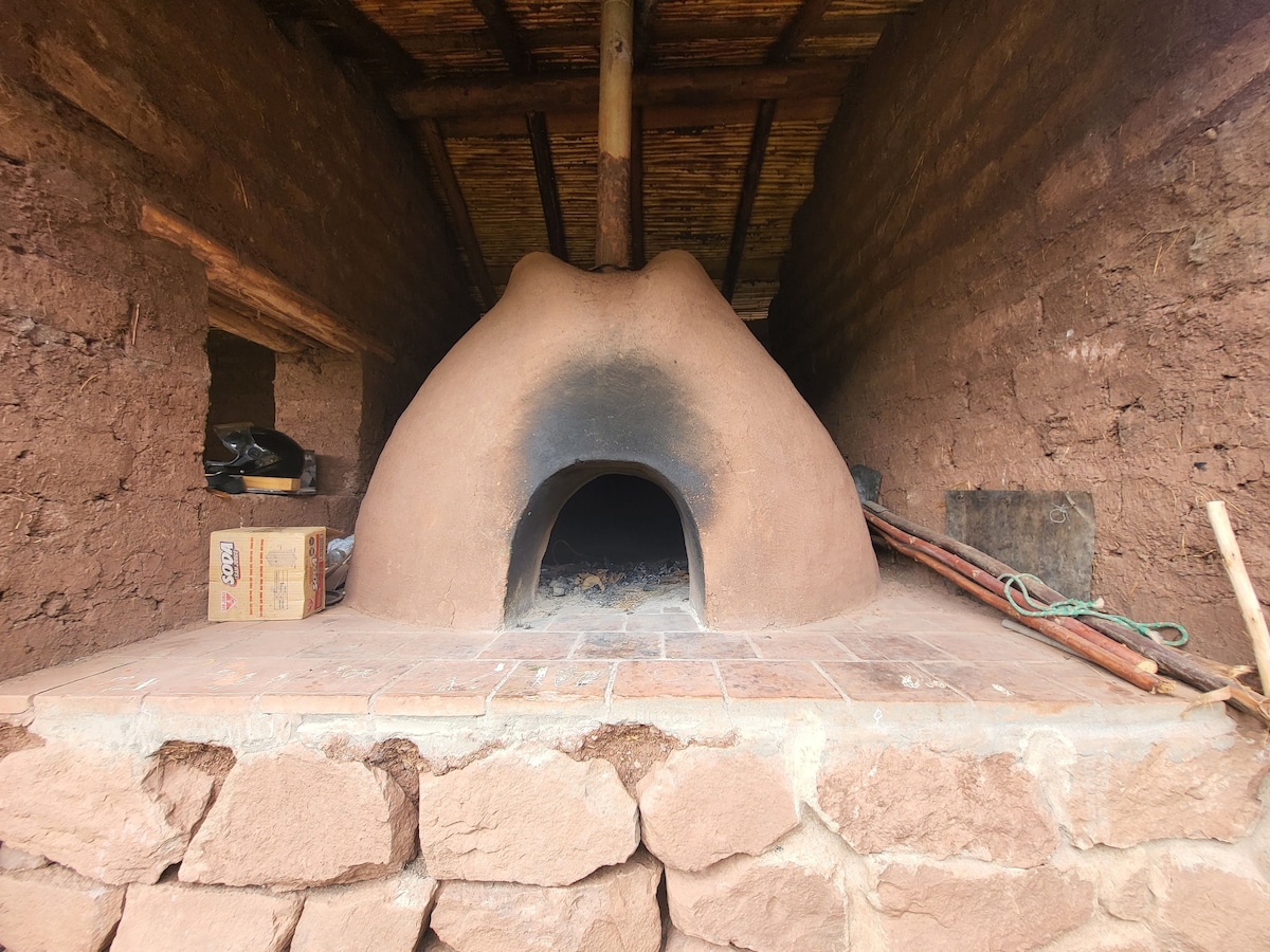 A traditional adobe brick oven is prominently featured, showcasing a rounded dome shape with a darkened entrance. The oven is set on a stone platform with tiled flooring, surrounded by rustic earth-toned walls and wooden beams above, evoking a sense of traditional craftsmanship.