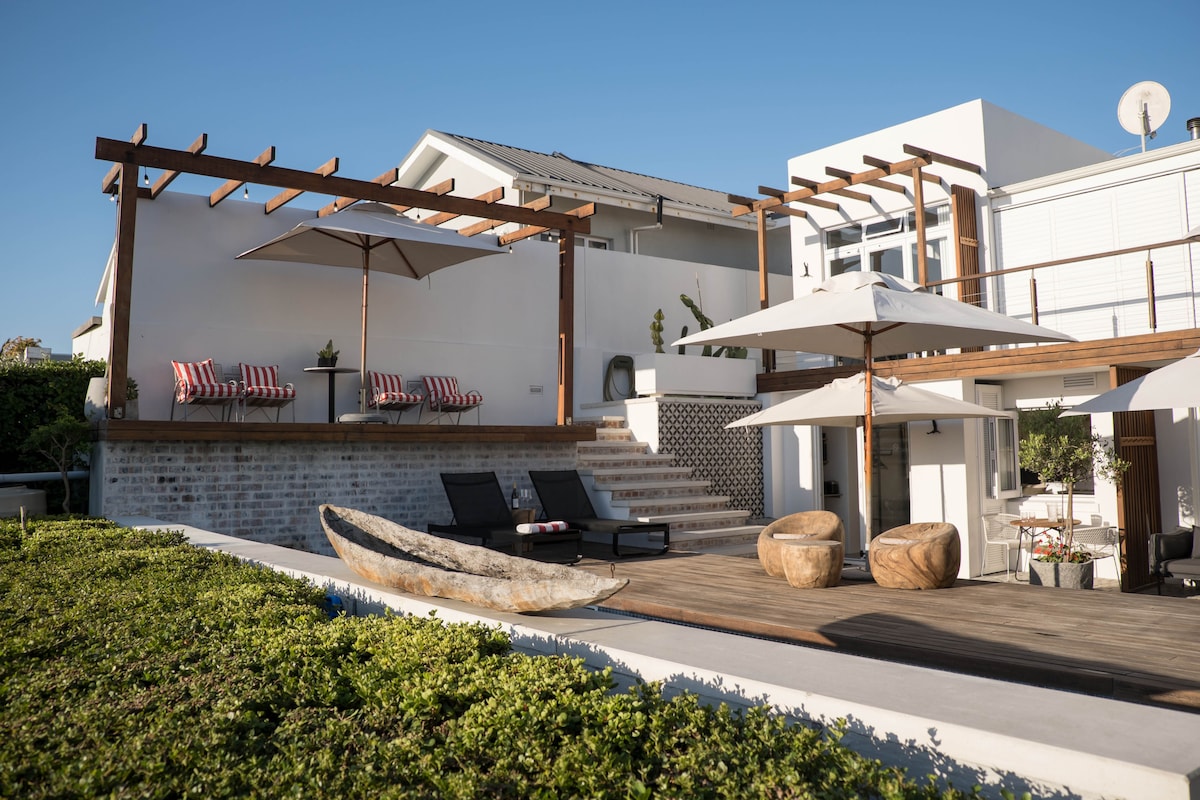 An outdoor deck is featured, highlighting two lounge chairs and a wooden boat accent. A pergola provides shade for seating, complete with two umbrellas. Steps lead to a well-maintained garden area, complemented by the modern architecture of the building in the background.