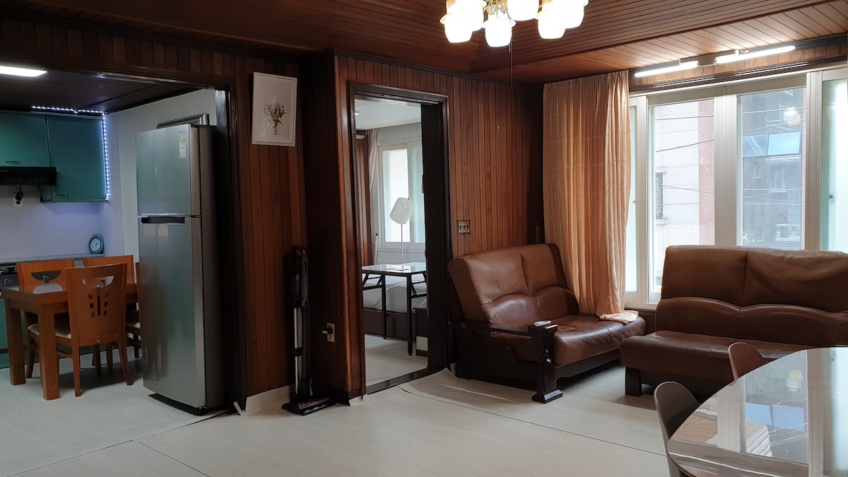 The image shows a spacious living area featuring wooden paneling and large windows that allow natural light to fill the room. A comfortable brown sofa set is positioned opposite a dining table, with a refrigerator and kitchen visible in the background.