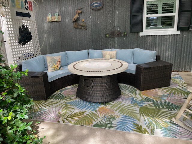 A circular seating arrangement is featured, upholstered in light blue fabric. The centerpiece is a round table with a fire pit at its center, surrounded by a textured outdoor rug displaying tropical patterns. The setting is shaded and creates a comfortable inviting space.