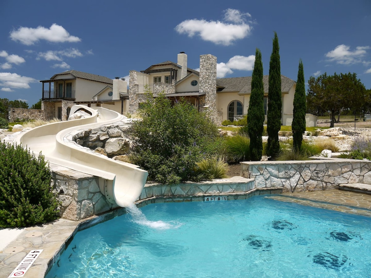 A clear blue swimming pool with a water slide curving down from a higher elevation. Lush greenery surrounds the area, complemented by elegant stonework. The background features a spacious building and tall, slender trees against a bright sky.