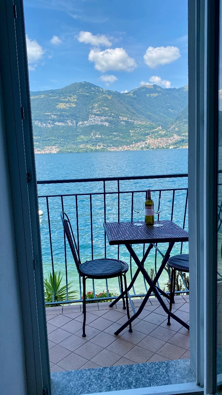 A balcony view is presented, featuring a small table with two chairs overlooking the serene lake, surrounded by greenery. The distant mountains and blue sky are clearly visible, creating a peaceful backdrop for relaxing moments.