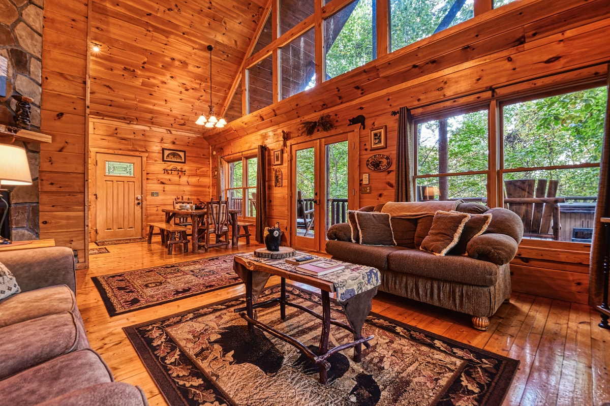 A spacious living room features high wooden ceilings and large windows, allowing natural light to fill the area. A comfortable sofa and a rustic coffee table are centered on a patterned rug, while a dining table is visible in the background, inviting gatherings.
