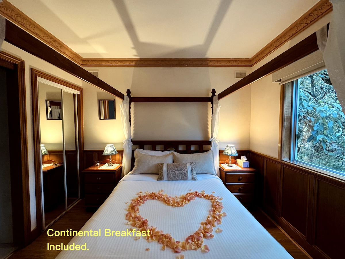 A well-appointed bedroom features a queen bed adorned with decorative petals arranged in a heart shape. Two bedside tables with lamps provide soft lighting. Natural light enters through a window, complemented by timber paneling along the walls, creating a warm ambiance.