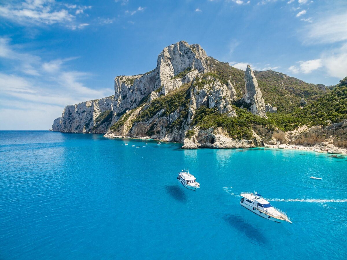 A serene coastline is depicted, featuring rocky cliffs rising from the vibrant blue waters. Two boats are seen navigating the calm sea, with the sunlight reflecting off the water's surface. Lush greenery covers the cliffs, enhancing the natural beauty of the landscape.