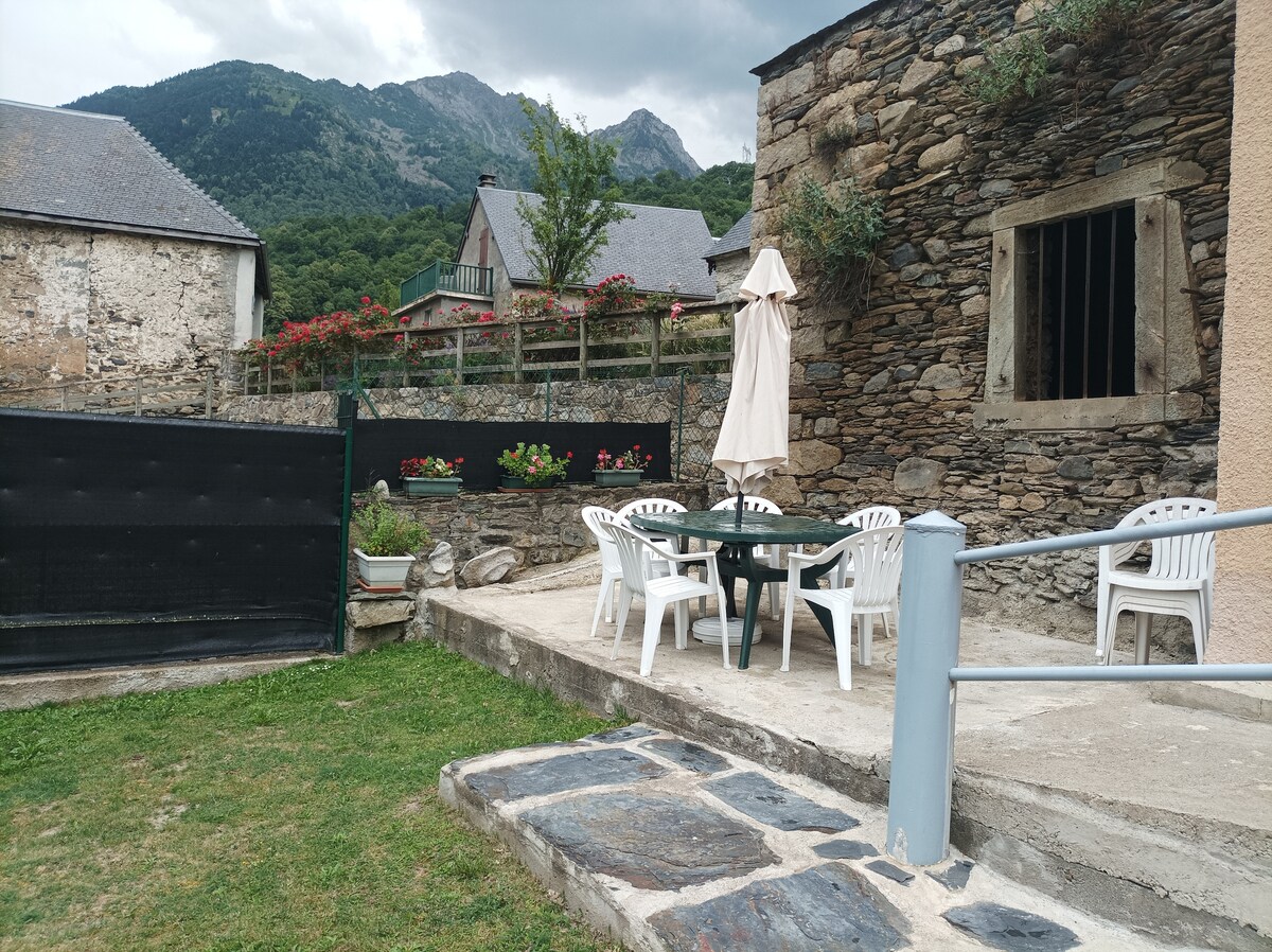 An outdoor terrace features a circular table surrounded by four white chairs, and an umbrella provides shade. The stone wall of the house adds texture, while a grassy area and mountain greenery are visible in the background, enhancing the natural ambiance.