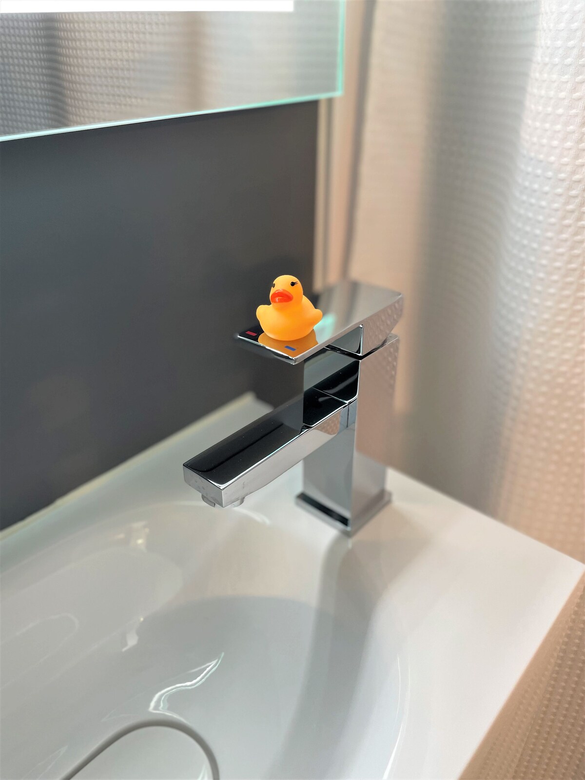 A modern bathroom faucet is shown with a sleek, minimalist design. A small yellow rubber duck rests on the faucet's edge, contrasting with the chrome finish. The sink features a clean, white basin, while the backdrop includes a textured wall that adds depth.