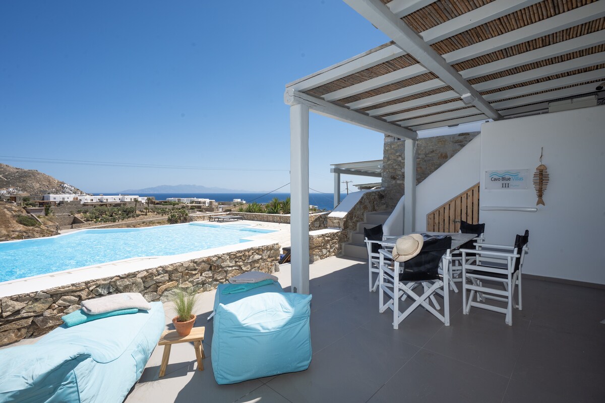 An outdoor terrace features a shaded area with a dining table and chairs facing a large swimming pool. Two bean bags are placed on one side, with a scenic view of the sea and landscape in the background.