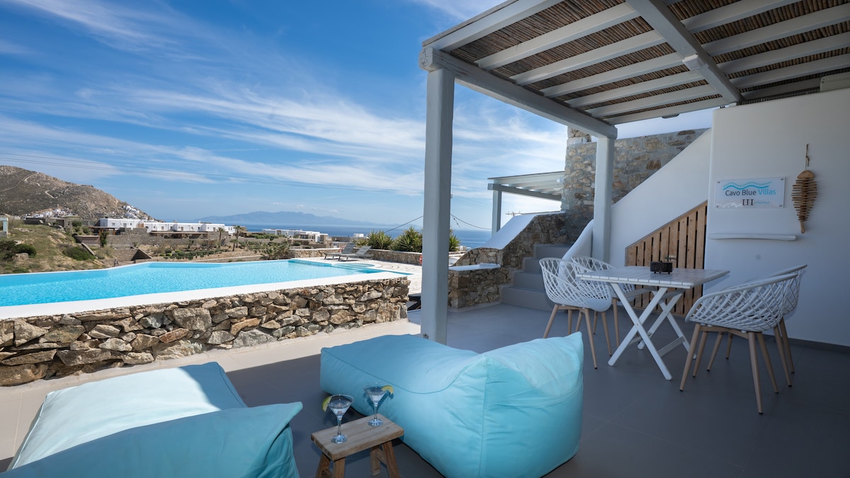 A terrace area is shown, featuring lightweight chairs and a table, with a stunning view of the large pool and surrounding landscape. The serene blue of the water contrasts with the natural stone walls, and inviting sunbeds offer a relaxed setting for outdoor enjoyment.
