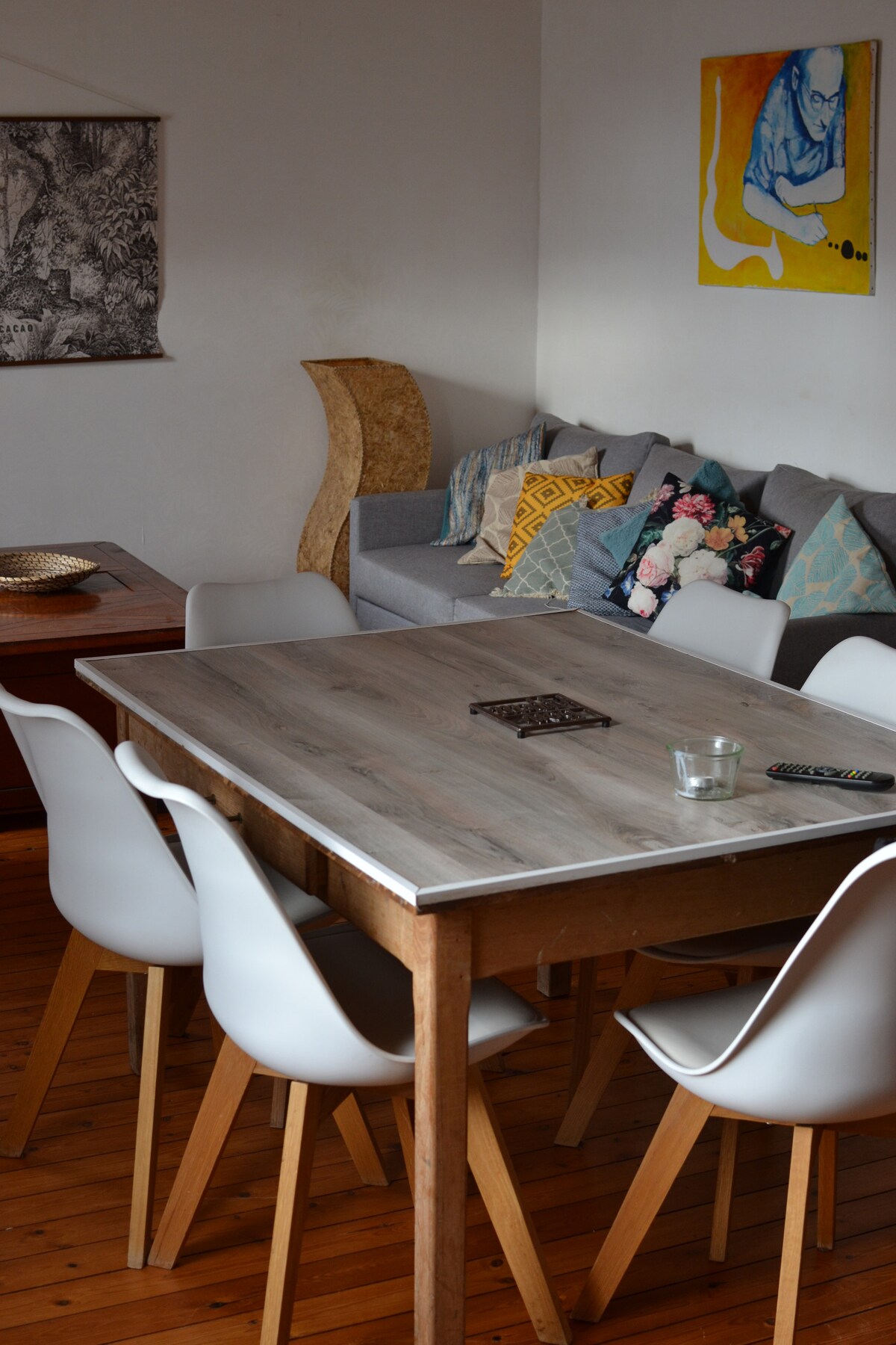 A dining table with a light wooden top is surrounded by six white chairs. A cozy seating area features a gray sofa adorned with colorful throw pillows. A piece of wall art adds a pop of color to the room, enhancing the overall aesthetic.