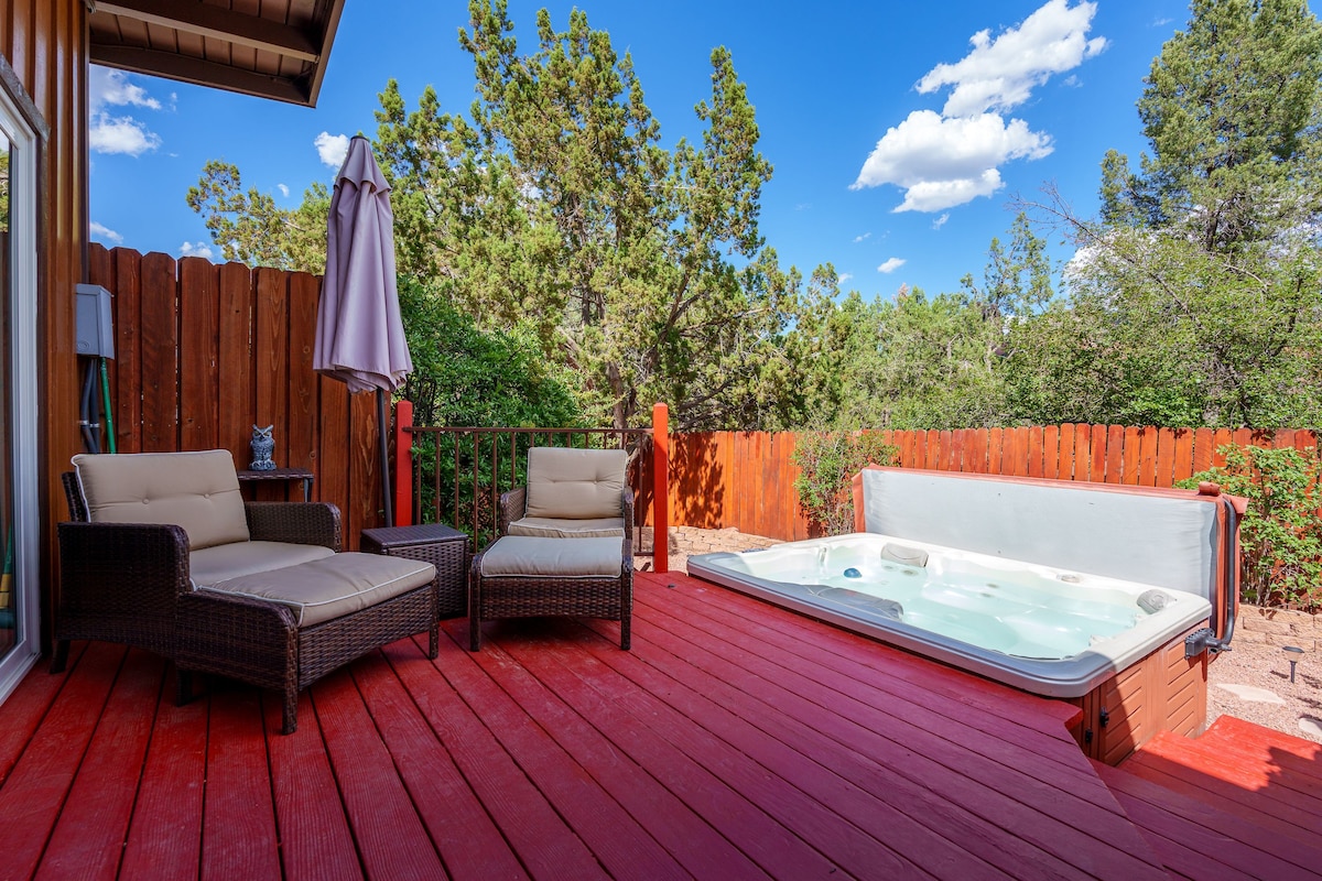 A spacious deck is presented with a hot tub and two cozy outdoor chairs. Rich wooden fencing provides privacy, while a large umbrella offers shade. Surrounding greenery adds to the natural ambiance, and the clear blue sky enhances the tranquil setting.