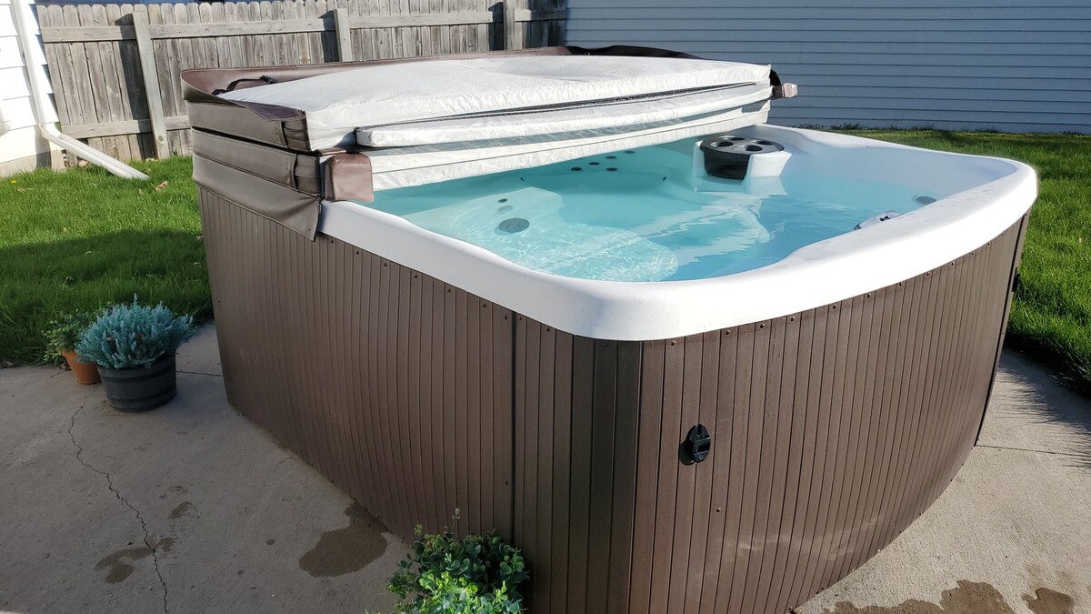 A hot tub with a light blue interior is positioned on a concrete patio, partially covered by a protective hard cover. Surrounding the hot tub, small potted plants add a touch of greenery, contrasting with the brown exterior of the tub.