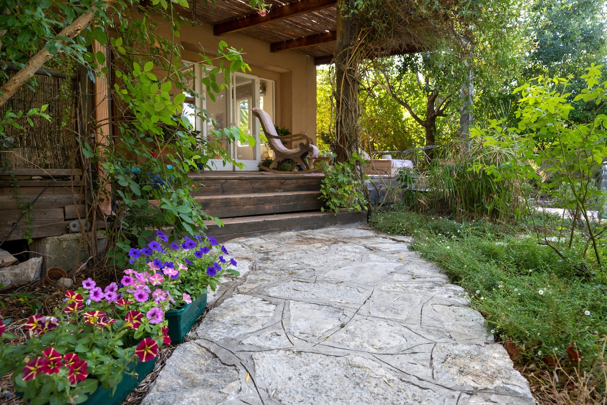 A stone pathway leads up to a cozy entrance, framed by vibrant flowers in various shades. Lush greenery surrounds the area, enhancing the natural setting. A wooden deck is visible, offering a welcoming outdoor space.