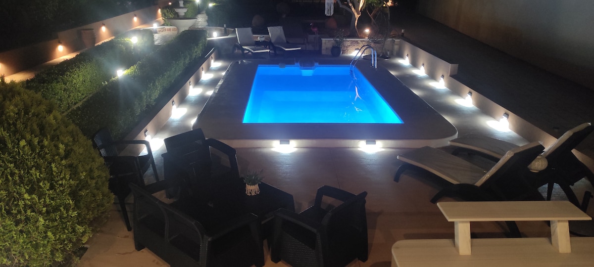 A well-lit outdoor space features a rectangular pool with vibrant blue water, surrounded by lounge chairs and small tables. Soft lighting outlines the pool area, enhancing the serene atmosphere, while neatly trimmed hedges create a sense of privacy.