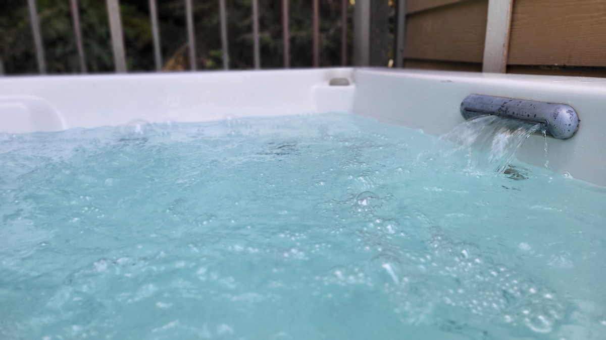 The hot tub is filled with clear, bubbling water, inviting relaxation. A smooth spout releases a gentle stream, creating ripples on the surface. The surrounding wooden fence adds privacy, allowing for a serene environment.