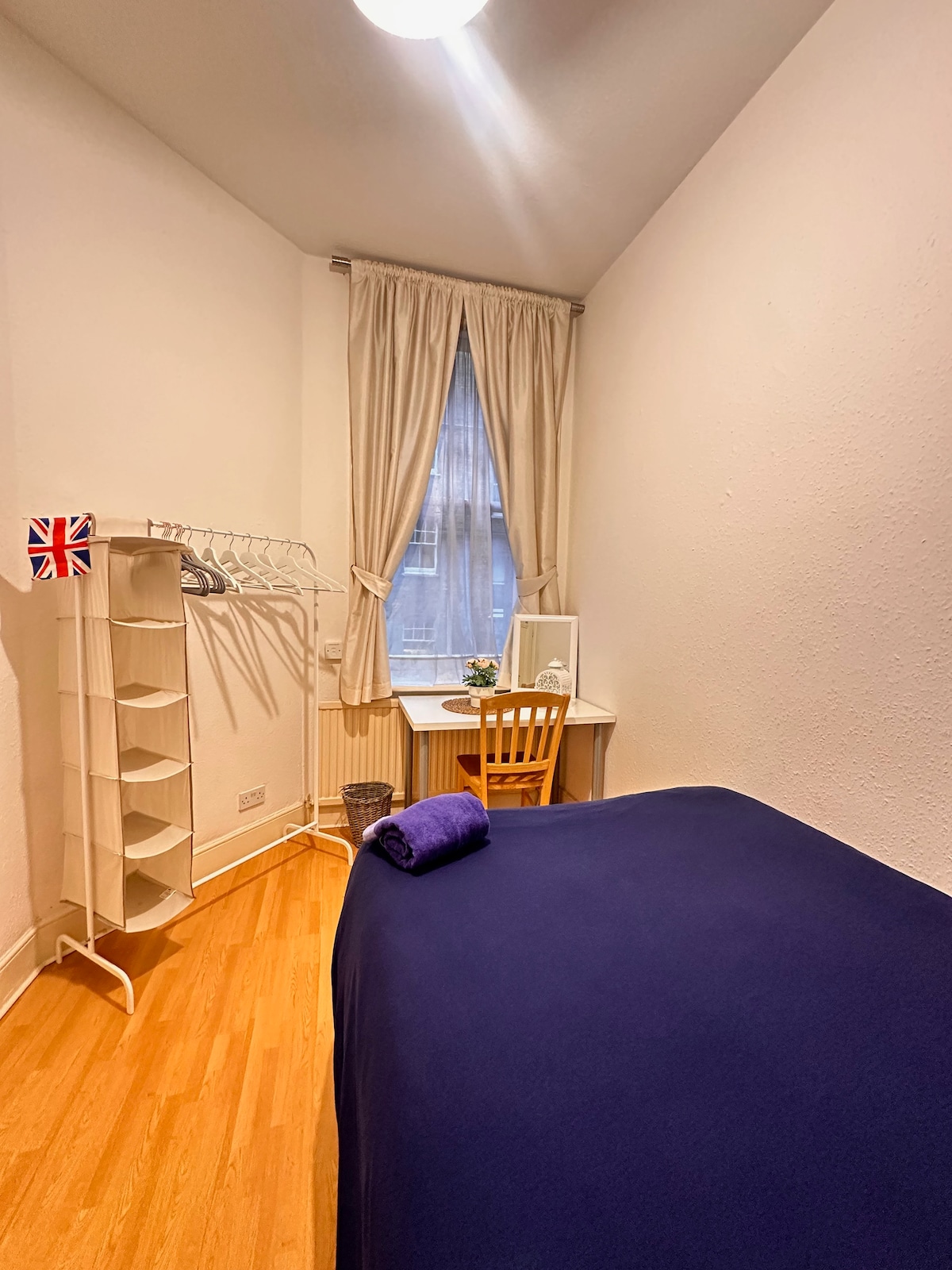 A cozy room features a spacious bed dressed in navy linens. A small desk with a chair is positioned near a window adorned with soft curtains. A clothes rack is placed nearby, providing additional storage. Light hardwood flooring complements the minimalist design.