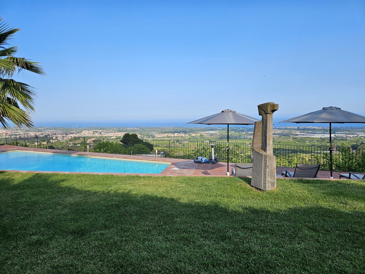 A serene outdoor space features a large swimming pool overlooking expansive views of the landscape and coastline. Two umbrellas provide shade over lounge chairs, while a stone sculpture complements the natural setting. Lush grass surrounds the pool area, enhancing the tranquil environment.