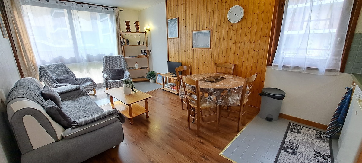 A living space features two armchairs and a sofa, complemented by a wooden dining table surrounded by chairs. Natural light enters through curtains on large windows, illuminating the wooden accent wall and the decor on shelves. A clock is mounted above a framed mirror.