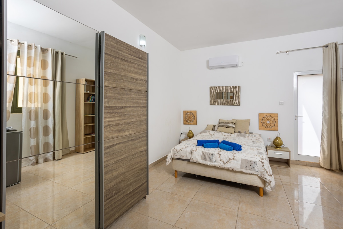 A spacious sleeping area features a large bed with decorative bedding, flanked by nightstands. A closet with mirrored doors adds to the room's brightness. Light enters through a door and window, while beige and cream tones create a calming atmosphere. Soft curtains frame the windows.