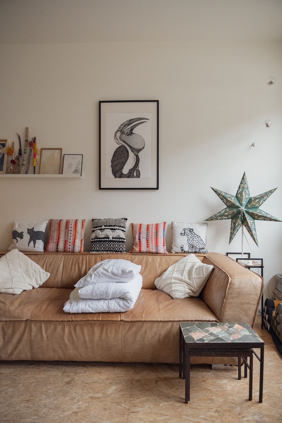 A cozy living area is furnished with a brown leather couch adorned with patterned cushions. A colorful star decoration hangs on the wall, and a small table, featuring a unique design, sits in front of the couch. Various art pieces are arranged on the nearby wall.