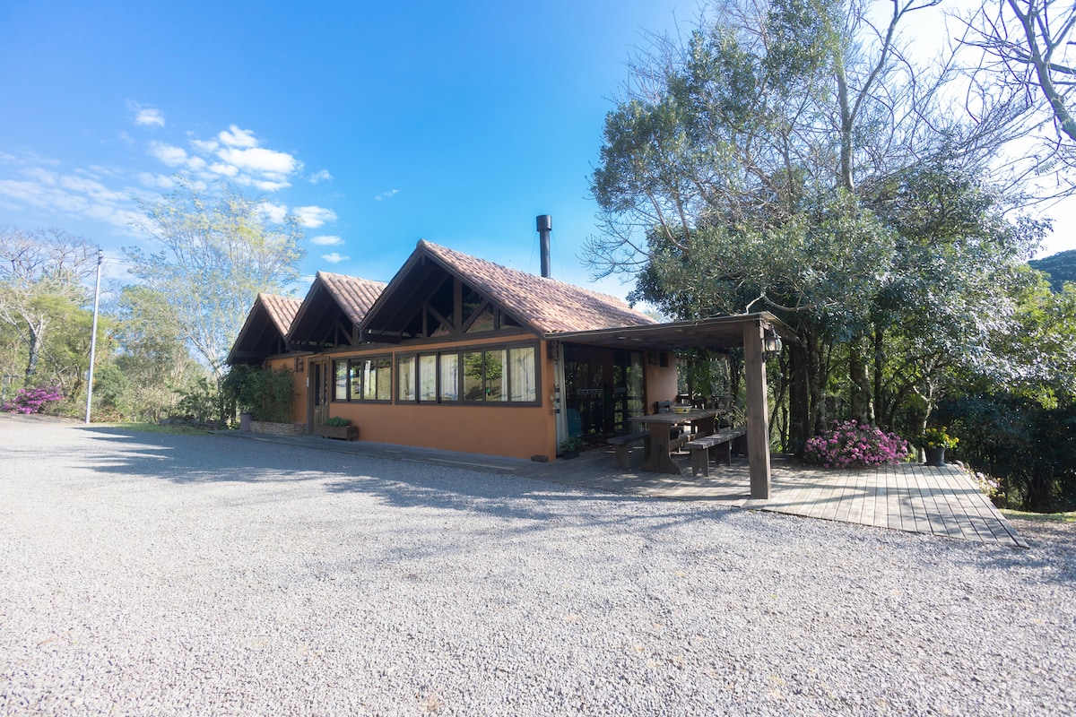 The exterior of a rustic house features a combination of warm earth tones and wooden accents. Large windows provide natural light to the interior. A spacious deck with outdoor seating is visible, surrounded by native vegetation and flowering plants, contributing to the serene rural setting.