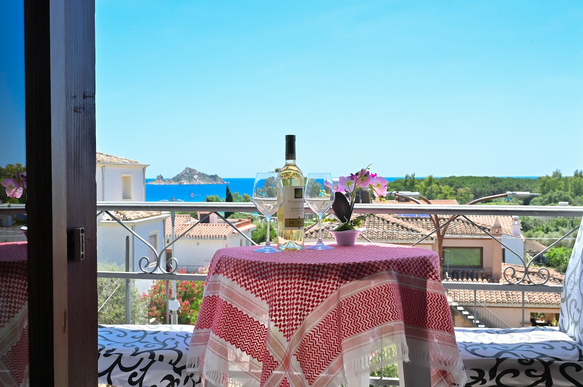 A small table is set on the balcony, featuring a bottle of wine and two glasses, fostering a relaxing ambiance. Floral decorations and a view of the sea and greenery are visible in the background under a clear blue sky.