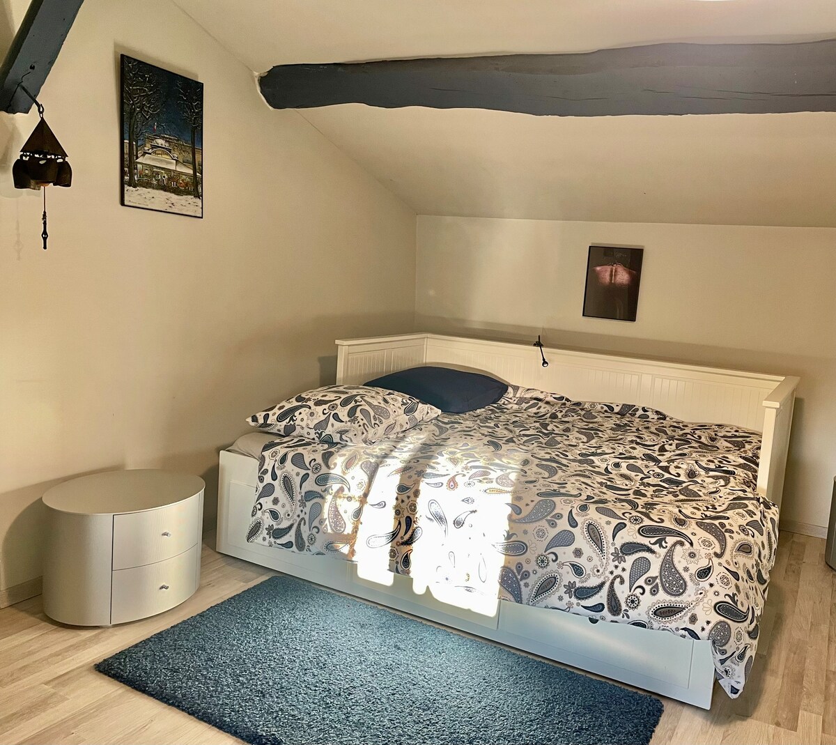 A spacious bedroom is highlighted by a large bed adorned with a patterned duvet. Natural light filters through a nearby window, illuminating the room. A round bedside table is positioned beside the bed on light wooden flooring, complemented by a soft area rug.