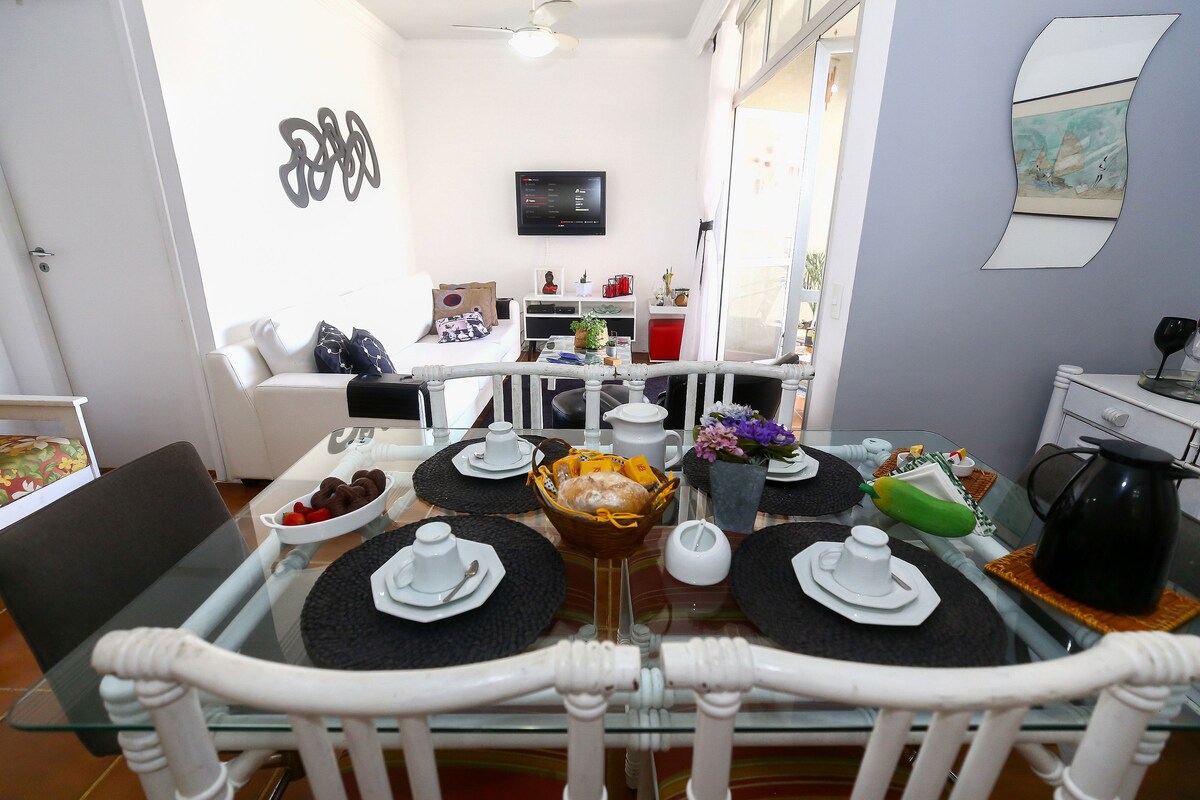 A dining area is set with white chairs surrounding a glass table. Plates and cups are arranged neatly on placemats. A cozy living space is visible in the background, featuring a sofa, TV, and decorative items. Natural light filters through nearby windows, creating an inviting ambiance.
