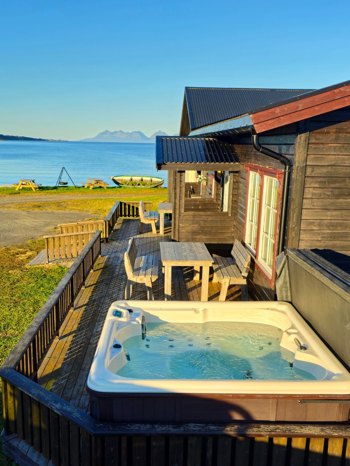 A wooden deck surrounds a hot tub, offering scenic views of the sea and distant mountains. Nearby, picnic tables are positioned on the lawn, inviting outdoor relaxation. The clear blue sky enhances the tranquil setting, with gentle waves visible at the water's edge.