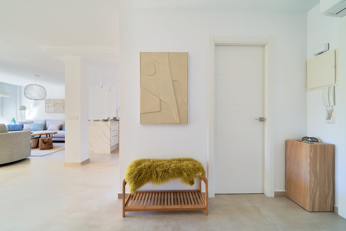 A well-lit entryway features a minimalist design with a light-colored wall and a textured artwork above a wooden bench. The bench is topped with a soft, green throw. A door is prominently positioned, leading to the closed space beyond.