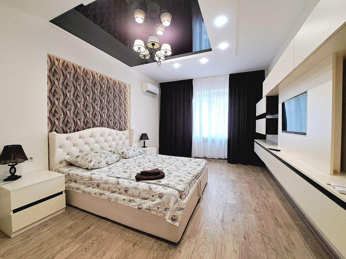 A spacious bedroom features a large bed with a tufted headboard and neutral bedding. Soft lighting is provided by a chandelier above, while a TV unit and a nightstand are positioned against the wall. Natural light fills the room through a window draped with sheer curtains.