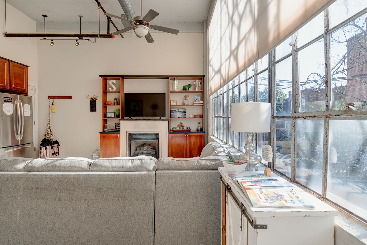 A spacious living area is featured, with a large sectional sofa facing a modern entertainment center. Large windows provide ample natural light, illuminating the warm wood cabinetry and decorative shelving. A simple side table with decor items adds to the inviting atmosphere.