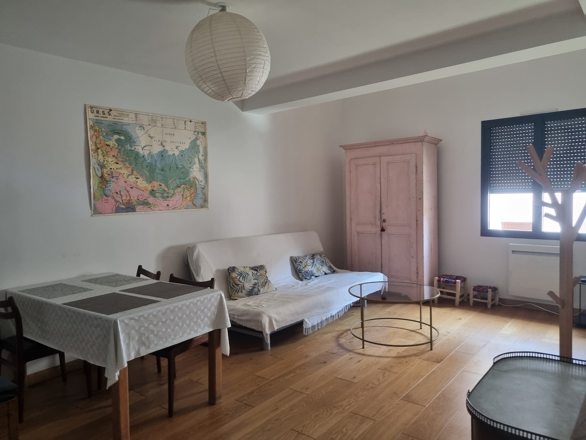A spacious living area features a white sofa bed, a round glass coffee table, and a dining table with seating for four. A soft pink wardrobe adds a touch of color, and a map adorns the wall, while natural light filters through the window with dark blinds.