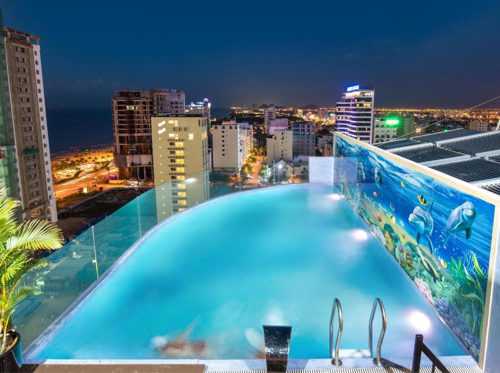 An infinity pool is visible against a city skyline illuminated by night lights. The water reflects the vibrant hues of adjacent murals, depicting marine life. The rooftop area is enclosed with glass panels, providing an unobstructed view of the surrounding landscape and nearby coastline.