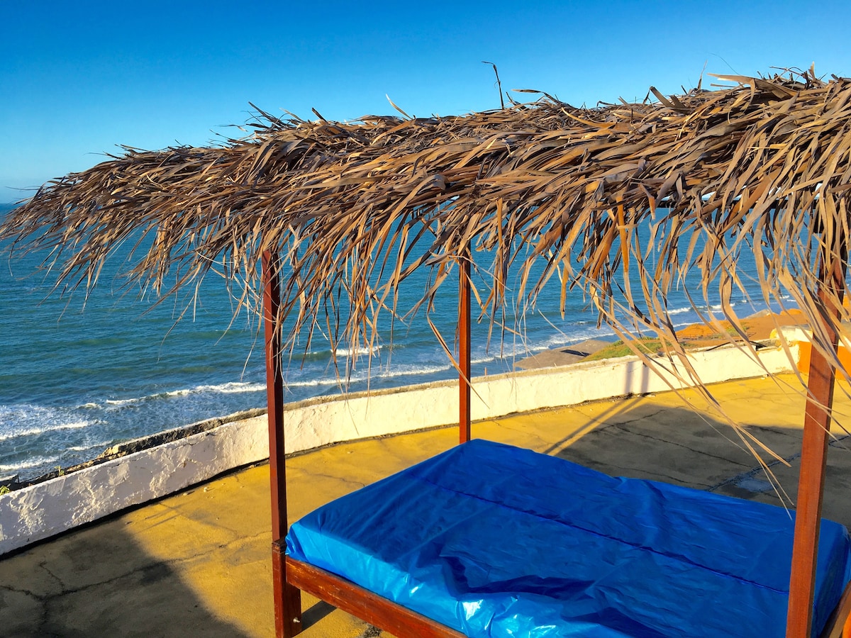 A shaded seating area is positioned on a rooftop terrace, offering views of the ocean. The structure features a thatched roof made of dried palm leaves, supported by wooden beams. A blue mat, resting on a flat surface, invites relaxation while enjoying the coastal scenery.