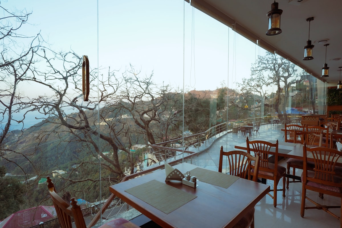 A dining area is presented with wooden tables and chairs, offering expansive glass walls that provide a view of the surrounding hills. The space is well-lit, with hanging lanterns and a partially covered terrace that allows for open-air dining.