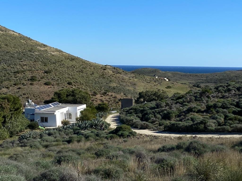 A white, eco-friendly house is nestled against a backdrop of rolling hills, surrounded by lush greenery. The distant blue sea is visible in the background, enhancing the natural beauty of the landscape. A dirt path leads to the property, reflecting the serene, remote location.