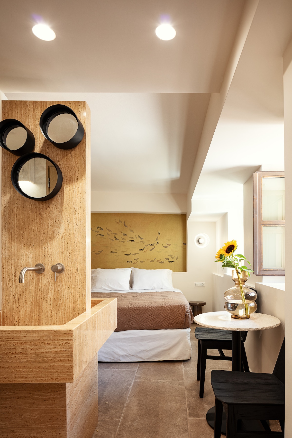 A spacious suite features a marble bathroom with a unique sink design, accompanied by three round mirrors. A cozy bed, adorned in neutral tones, is visible in the background. A table with two chairs is positioned near the window, enhancing the suite's airy ambiance.